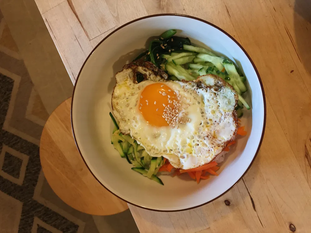 Bibimbap Bowl of bibimbap topped with a fried egg, sesame seeds, cucumber, carrot, and spinach