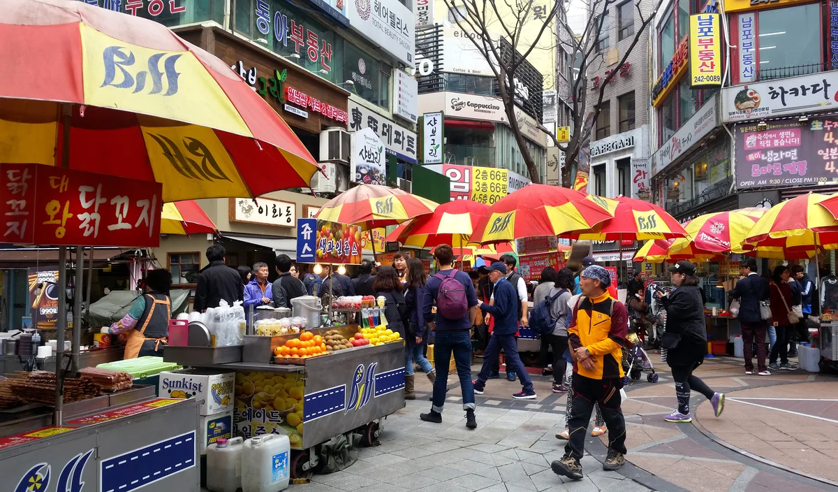 Street food stalls under red and yellow umbrellas at BIFF Square in Nampo-dong, Busan, with crowds of pedestrians