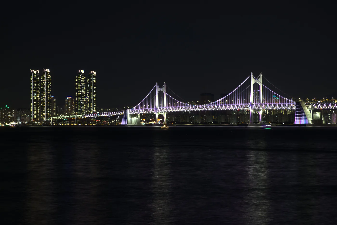Gwangan Bridge illuminated at night with the Busan skyline behind it, seen from across the water