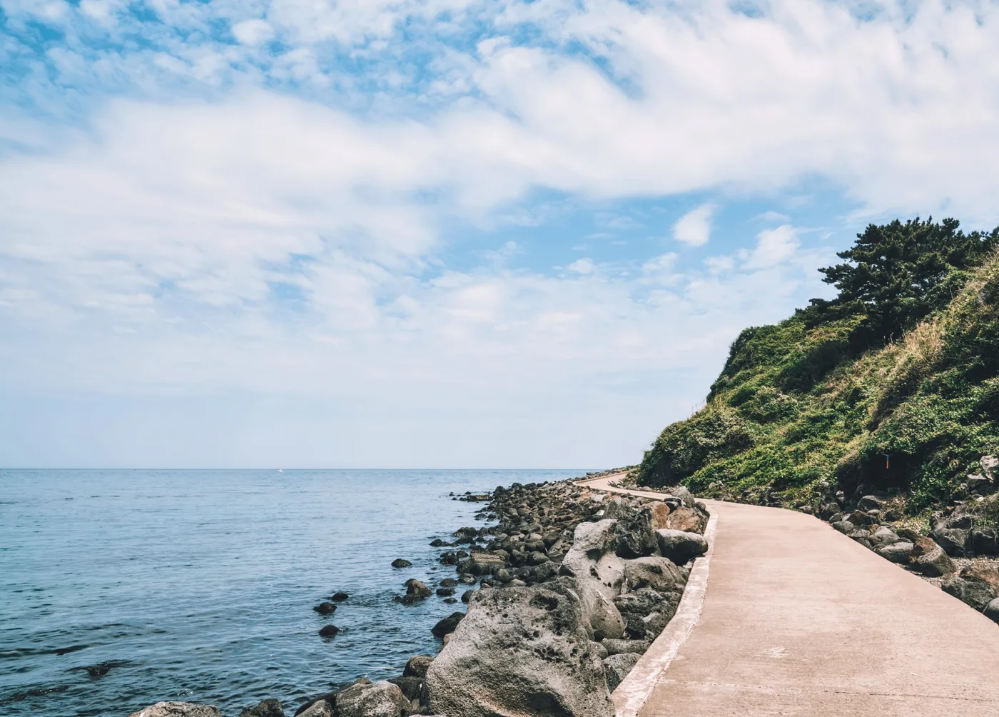 A paved coastal walking path along the Jeju Olle Trail, following a rocky shoreline with the sea on one side