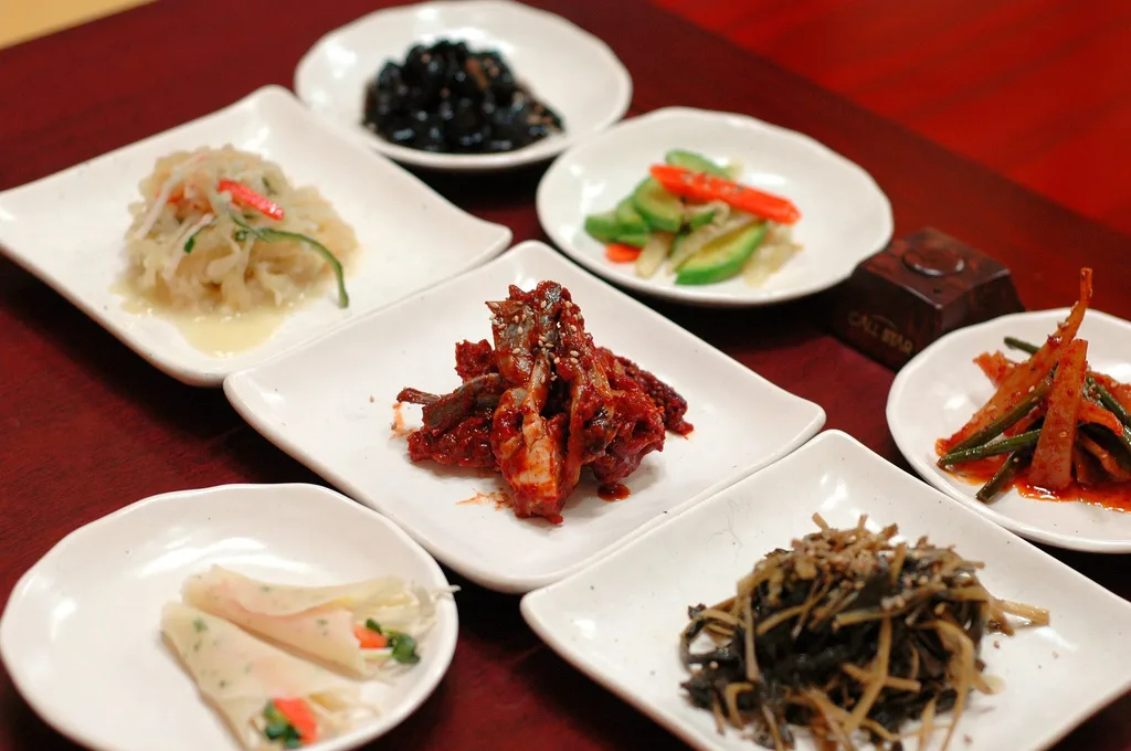 An assortment of small Korean banchan side dishes served on white plates, including seasoned vegetables, pickles, and spicy braised anchovies