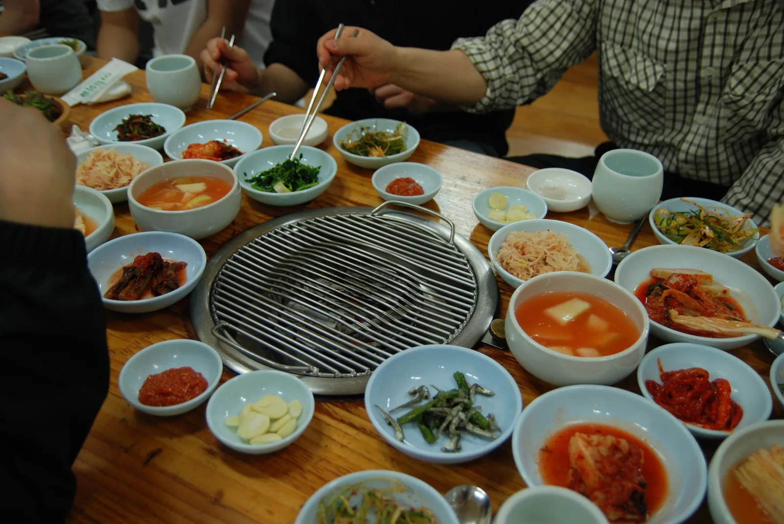 A tabletop Korean BBQ grill surrounded by a full spread of banchan side dishes, with diners' chopsticks reaching in
